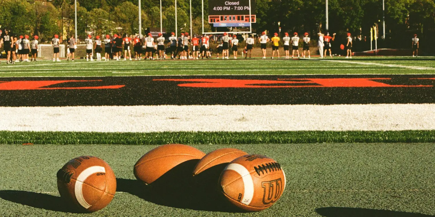 Footballs resting on the turf while a college team warms up on the field behind them