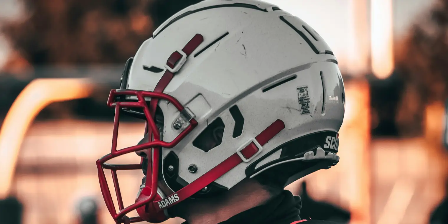 A college football player in a white helmet with a red face mask looks out over the field at sunset