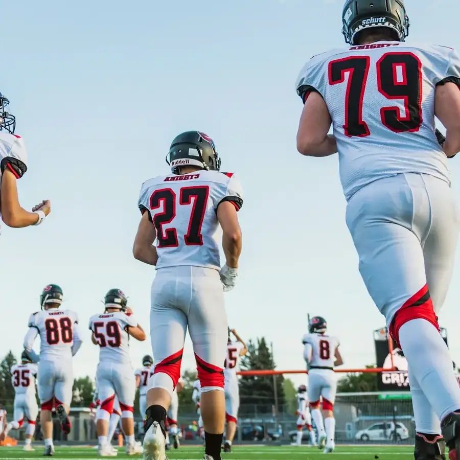High school football players in white jerseys jogging onto the field before a game
