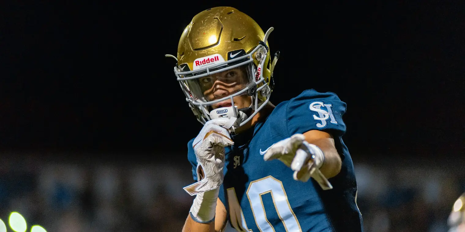 High school football player in a gold helmet and blue jersey pointing toward the camera under stadium lights